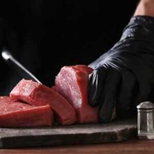Person's hands with black gloves holding a chef's knife, cutting slices of steak.